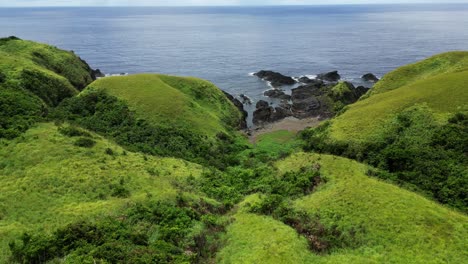 Gentle-sloping-green-lush-vegetated-hillsides-lead-to-basalt-rocks-on-beach,-philippines