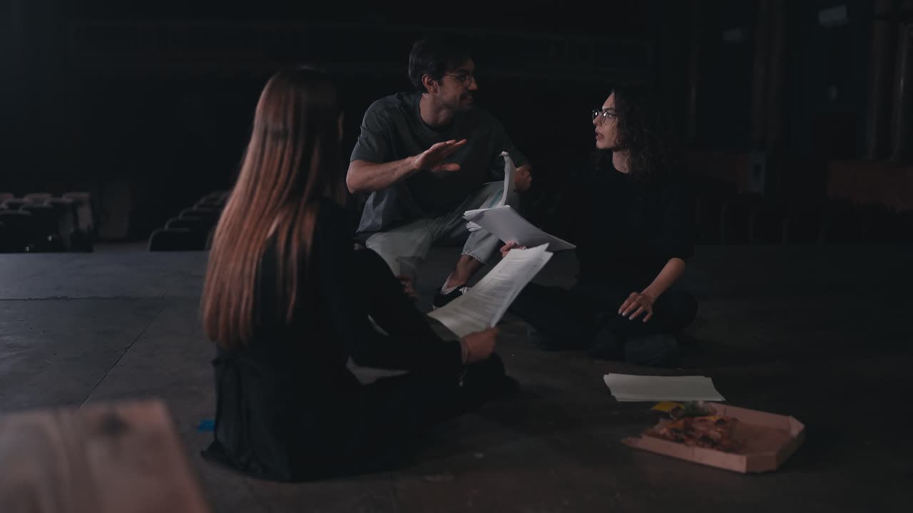 Two female actresses in black suits sit on stage and communicate holding in their hands the text of their characters and the script of the play while their director runs up to them and points out how to play correctly