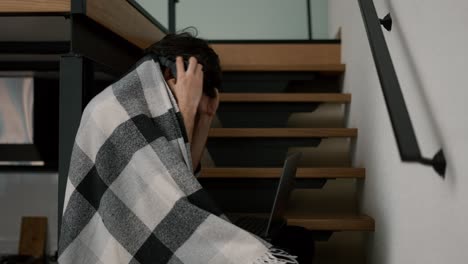 Bearded-man-with-laptop,-sitting-on-wooden-stairs-at-home-in-plaid-making-music