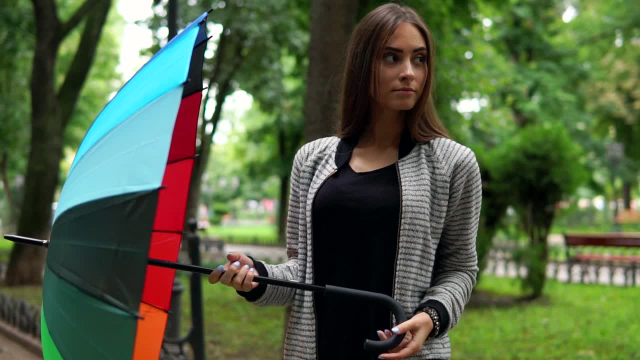 retrato de una joven atractiva mujer morena girando su colorido paraguas en el parque de la ciudad y mirando a la cámara