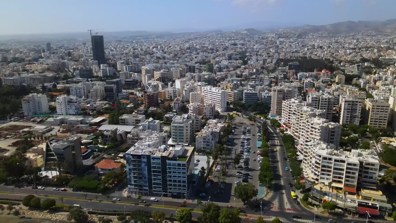 Aerial footage over urban Durrës, Albania, showcasing a towering skyscraper amidst dense cityscape, framed by vast mountains under a clear blue sky, amplifying urban density