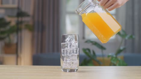 orange juice being poured into a glass with ice cubes on the table at home