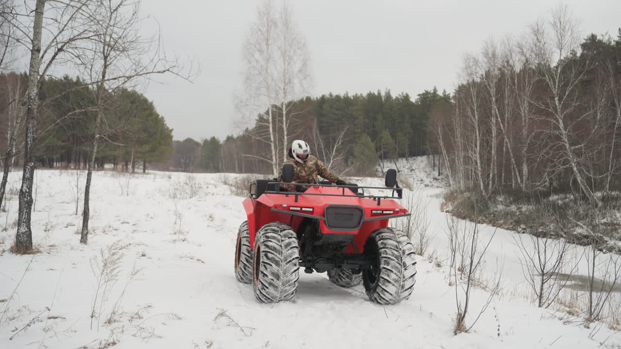 ATV rosso parcheggiato in una radura innevata con pilota in tenuta mimetica e casco. Vista frontale del quad rosso su tracce di neve, betulle e pini che costeggiano un prato ghiacciato, sotto un cielo coperto e tranquillo. Primo piano di pneumatici e motore.
