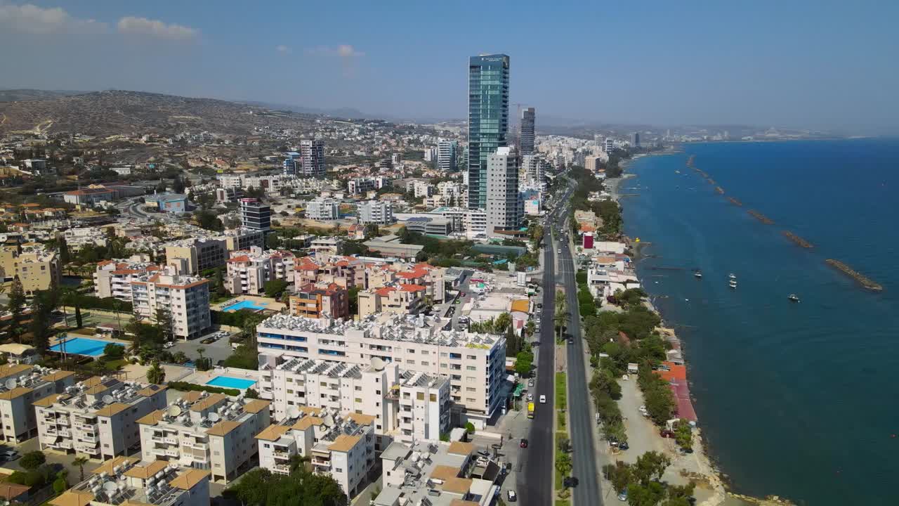 Breathtaking aerial footage of Sarandë, Albania, showcasing the urban coastline with a prominent skyscraper, winding road, and picturesque hilly backdrop on a clear, sunny day