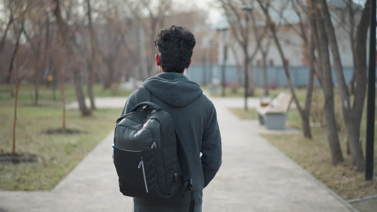 Young man with black hair seen from behind walking along paved park path wearing dark hoodie and carrying large black backpack, surrounded by bare trees and benches in quiet urban environment