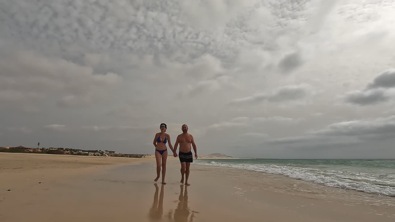 Romantic couple walks hand in hand along peaceful, sandy beach of Boa Vista island with cloudy sky overhead, creating serene and intimate moment by the ocean, Cape Verde. Slow motion
