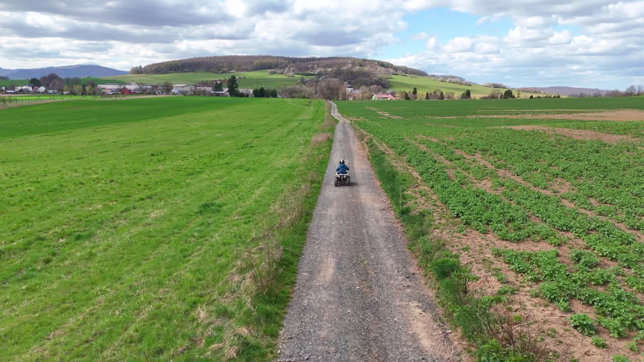 Vista aerea che segue un bambino che guida un quad lungo una strada sterrata diritta attraverso campi verdi aperti, paesaggio rurale pacifico con ampi scenari naturali.