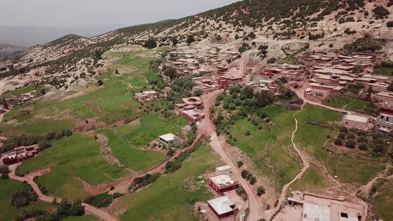 Toma aérea amplia que captura un extenso pueblo bereber tradicional construido en las escarpadas laderas del Alto Atlas. El vídeo destaca la mezcla de arquitectura de tierra roja y campos verdes aterrazados.