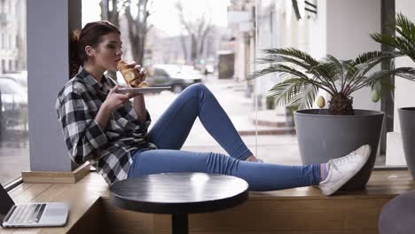 a brunette girl in a plaid shirt and jeans sits with outstretched legs by the window and enjoys her croissant. smiling. blurred street background