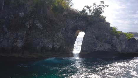 Waves-crashing-through-a-hole-in-rocky-escarpment-on-a-sunny-day-in-Crystal-bay,-Nusa-Penida,-Indonesia