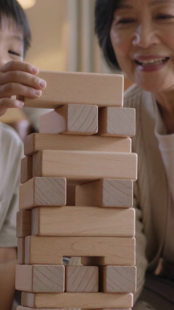 Vertical video: Reaching child placing wooden block balancing tower on carpet, senior letting go