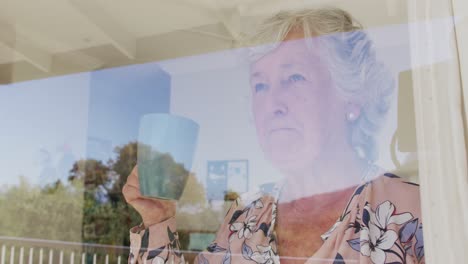 caucasian senior woman drinking coffee while looking through the window at home