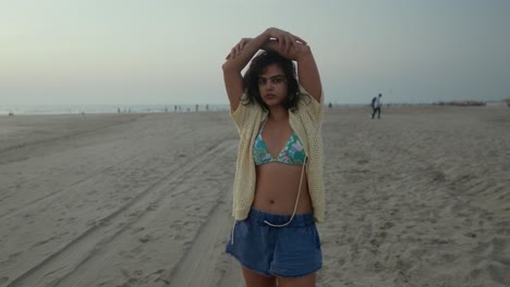 young woman in a bikini top and open cardigan at the beach during dusk, arms raised, casual and relaxed