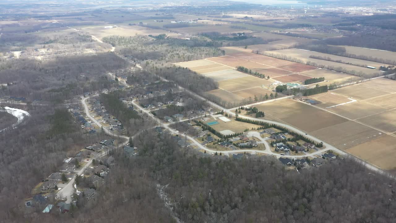 Volo ad alta quota sopra un quartiere periferico situato tra una foresta spoglia e vasti campi agricoli, che mostra il contrasto tra la vita residenziale e la campagna rurale