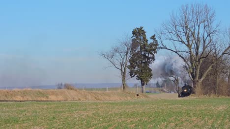 a restored steam passenger train passes by blowing white and gray smoke, traveling thru rural countryside on a sunny winter day