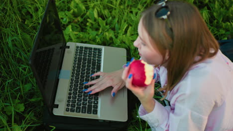 russian lady lying on grass with glasses on head typing on laptop with right hand while holding partially eaten apple in left hand, relaxed outdoor setting surrounded by vibrant greenery