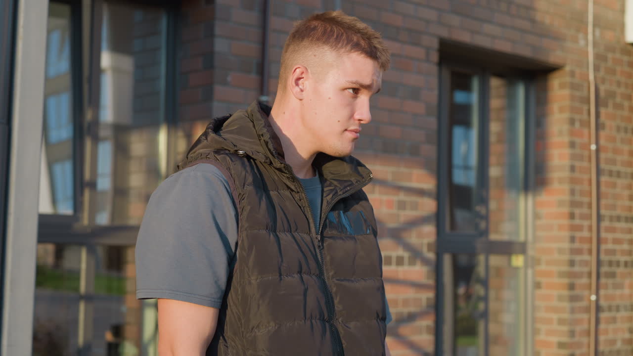young man in black puffer vest and grey shirt standing near brick building holding cigarette pack in hand while looking up thoughtfully