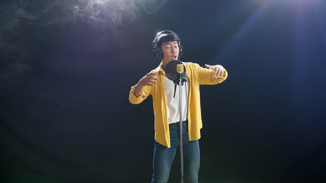 헤드폰을 들고 있는 젊은 아시아 소년이 색 연기가 있는 검은색 배경에 있는 콘덴서 마이크에 을 치고 있다.