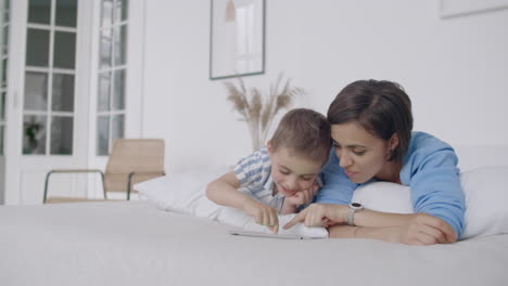 madre e hijo usando tableta digital en el dormitorio en casa. vista frontal de madre y hijo caucásicos felices usando tableta virtual en el dormitorio en casa