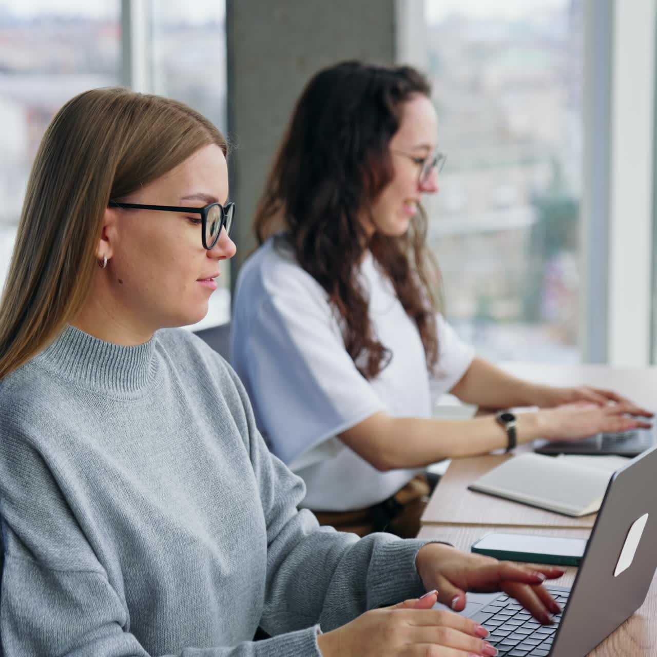 Two positive ladies work at laptops in the office. Women wearing computer glasses type and talk
