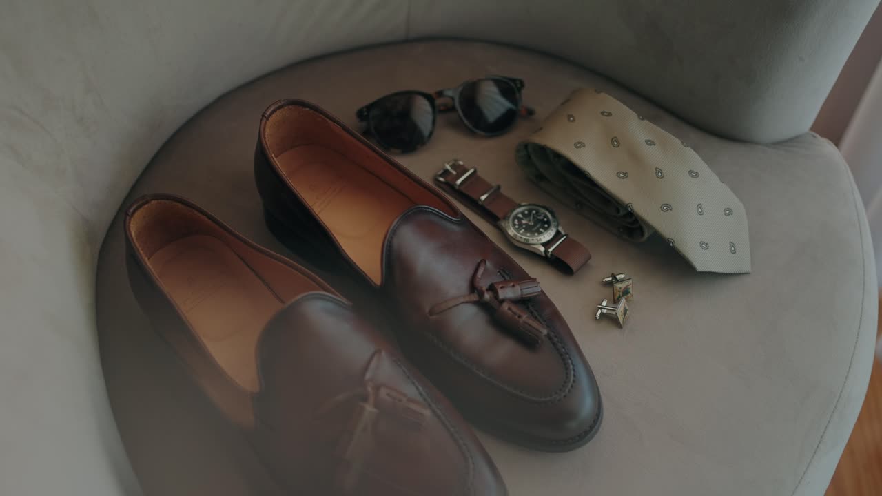 flat lay of brown loafers sunglasses watch cufflinks and tie arranged on a cushioned chair