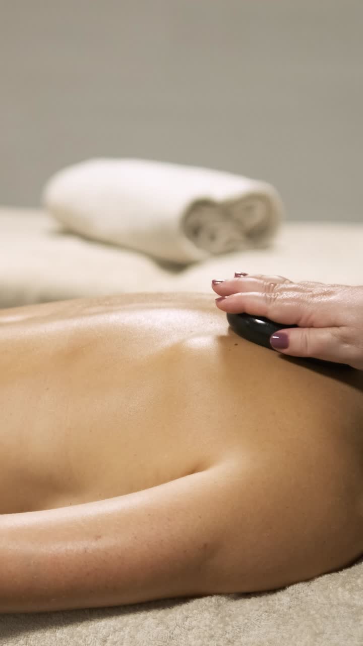 Masseuse delivering a relaxing hot stone massage treatment on a client's back. Vertical