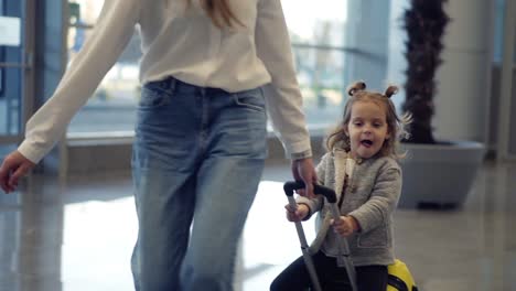 Airport-terminal,-young-unrecognizable-mother-riding-her-cute-daughter-on-a-small-yellow-suitcase.-Mom-with-yellow-suitcase-and-daughter-are-having-fun-before-their-departure.-Close-up