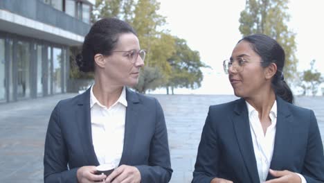 smiling female business colleagues walking at office building, drinking coffee and talking together