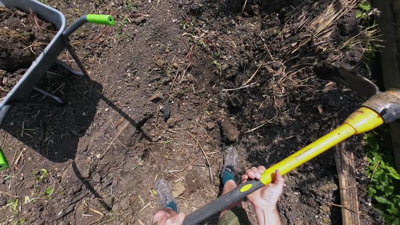 POV from a man using a pick axe to dig out a planter of soil in a garden on a sunny day