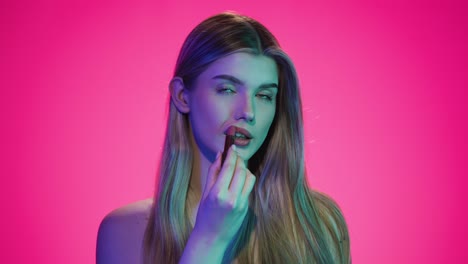 Medium-static-shot-of-a-pretty-young-woman-or-model-looking-sexy-and-seductive-into-the-camera-getting-ready-for-club-night-while-applying-her-red-lipstick-in-front-of-pink-background-in-slow-motion