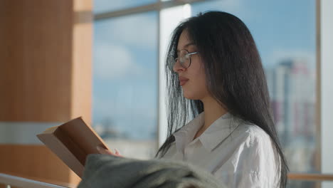 focused lady with long black hair and glasses flips page while reading book, resting beside coat on wooden rail in modern indoor space with sunlight and blurred urban cityscape through glass windows