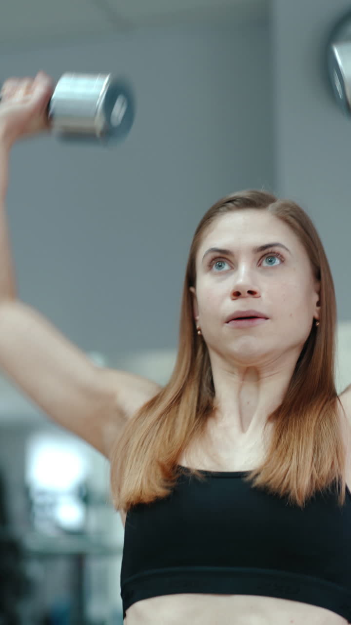 Pretty woman in black top raises up dumbbells on the background of the gym. Exercises for biceps. Close-up Vertical video