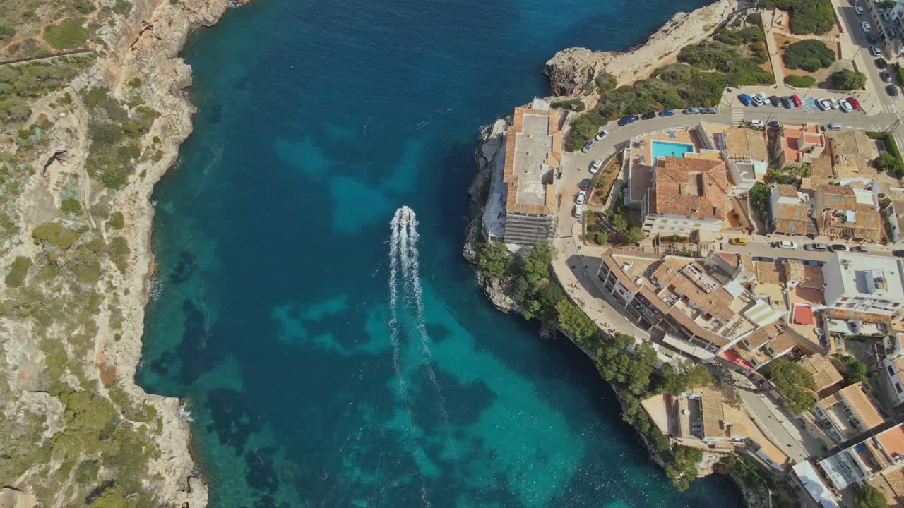 A drone captures a stunning view of a Mediterranean coastal town with terracotta-roofed buildings. A small boat sails through the clear turquoise bay, leaving a white wake behind.