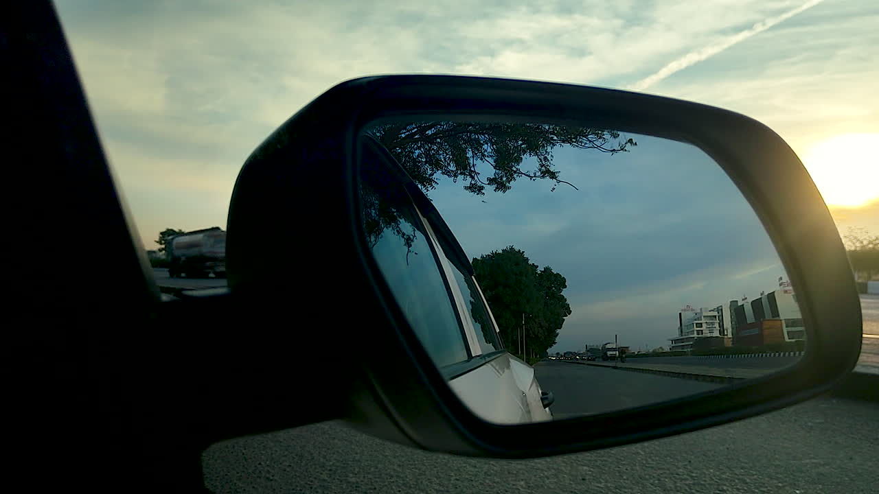 Specchietto laterale dell'auto che riflette strada e traffico autostradale sotto il cielo serale in India, a rappresentare il concetto di viaggio, spostamento, trasporto, libertà e stile di vita moderno