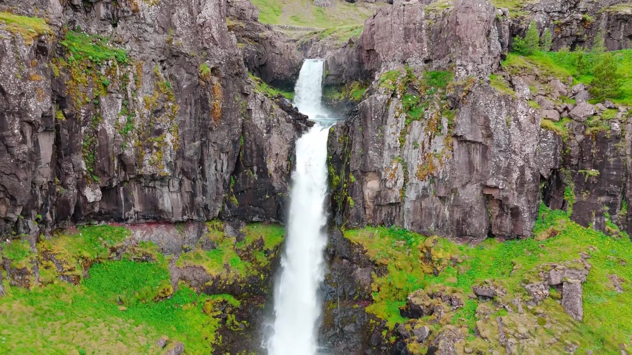 Et fantastisk luftfoto af Snadalsfoss-vandfaldet på Island, der viser vand, der fosser ned gennem barske basaltklipper og frodigt grønt mos. Ideel til rejse- og bæredygtighedsindhold.