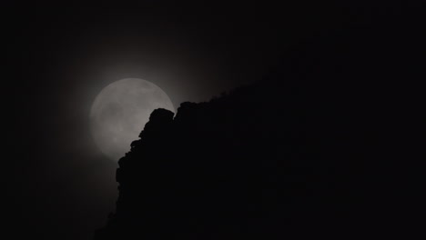 a full moon rises behind clouds and a rocky mountain foreground