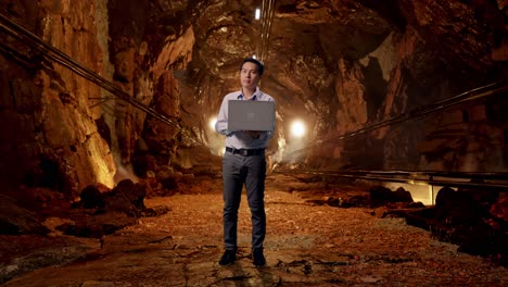 Full Body Of An Asian Male Professional Worker With His Laptop In Underground Mine Tunnel, He Observes By Looking Around Before He Come To Concentrating With His Laptop