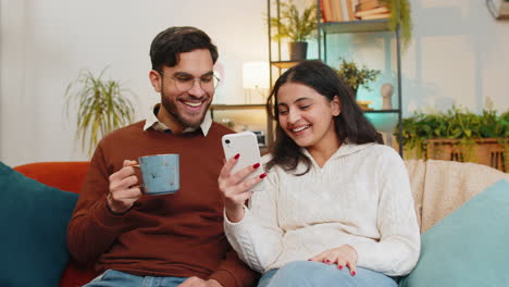 Happy-girlfriend-boyfriend-couple-browse-social-media-applications-together-on-smartphone-at-home