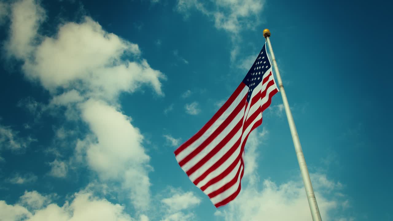 Vista dal basso della bandiera americana che sventola su un'alta asta contro un cielo azzurro brillante con nuvole bianche, simbolo patriottico degli Stati Uniti di libertà e orgoglio nazionale