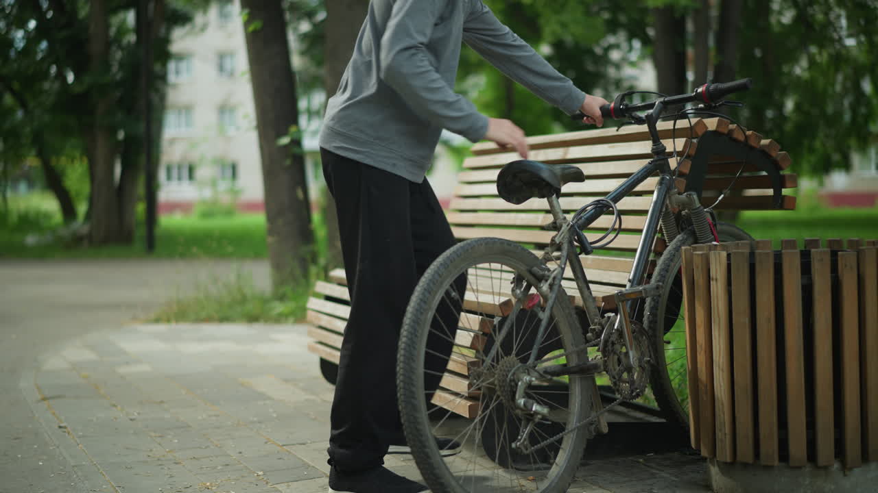 어린 소년은 평온한 공원에 자전거를 타고 가서 나무 벤치와 인근 구조물 사이에 주차합니다. 그는 자전거가 벤치에 앉을 때, 초록색과 나무로 둘러싸여 있습니다.