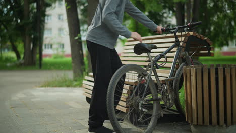 young boy rides his bicycle into a peaceful park and parks it between a wooden bench and a nearby structure, he lets the bicycle rest on the bench as he sits down, surrounded by greenery and trees