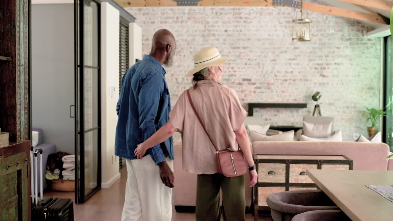 Diverse couple arriving in living area, senior man pulling cases, woman in straw hat exploring