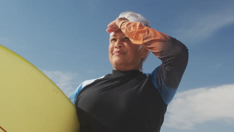 Happy-senior-hispanic-woman-standing-on-beach-with-surfboard