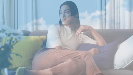 animation of sky with clouds over biracial woman sitting on sofa