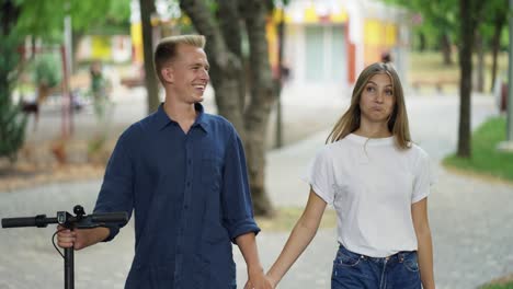 The-guy-and-the-girl-are-walking-in-the-park,-talking.-The-guy-is-holding-a-scooter-in-his-hand.-Happy-walk.-romantic-date