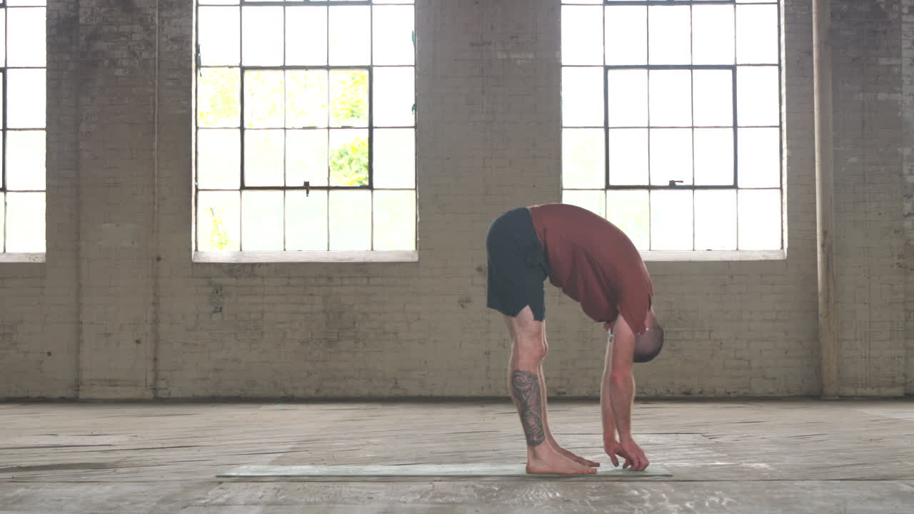 Man in an industrial warehouse practicing yoga, the pose of forward fold, reaching for his toes.