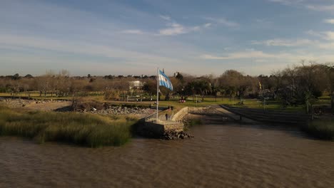 Aerial-flight-towards-Argentinian-flag-waving-on-river-coastline-during-sunset---Park-with-trees-in-background