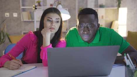 Focused-and-serious-working-multiethnic-married-couple-using-laptop-together-in-home-office.