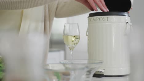 midsection of senior caucasian woman in kitchen, composting vegetable scraps and drinking white wine