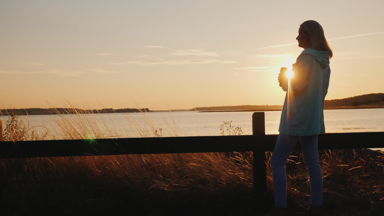 외로운 여자는 일몰을 바라보며 울타리 옆에 홀로 서 있는 호수 위 캔에서 맥주를 마신다